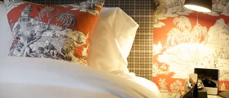 A neatly made bed with a pillow featuring a traditional design, a bedside table with a telephone and a lamp, and a wall with a patterned mural.