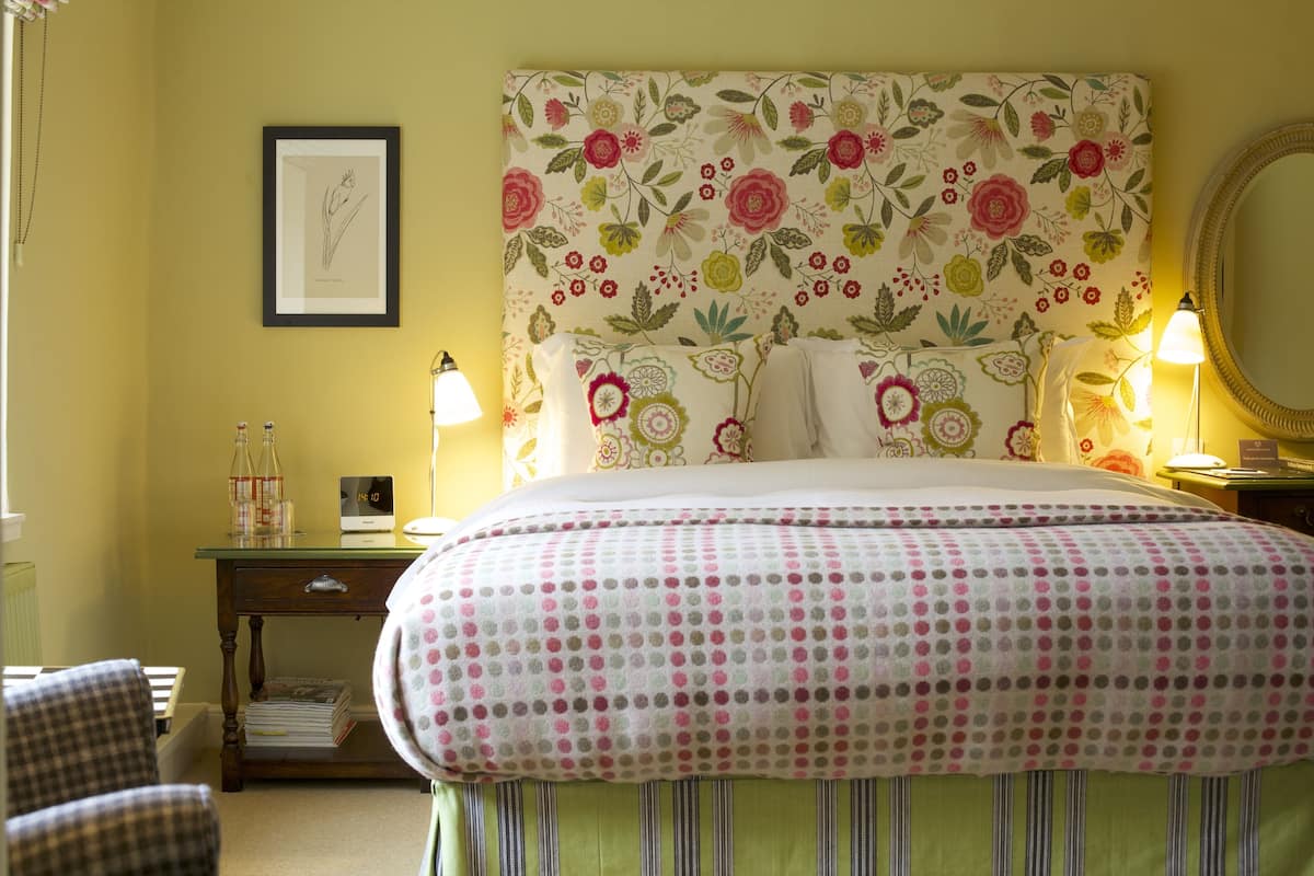 A neatly arranged hotel room with a floral-patterned headboard, a bed with a polka dot and striped bedspread, a bedside table with a lamp, and a framed botanical illustration on the wall.