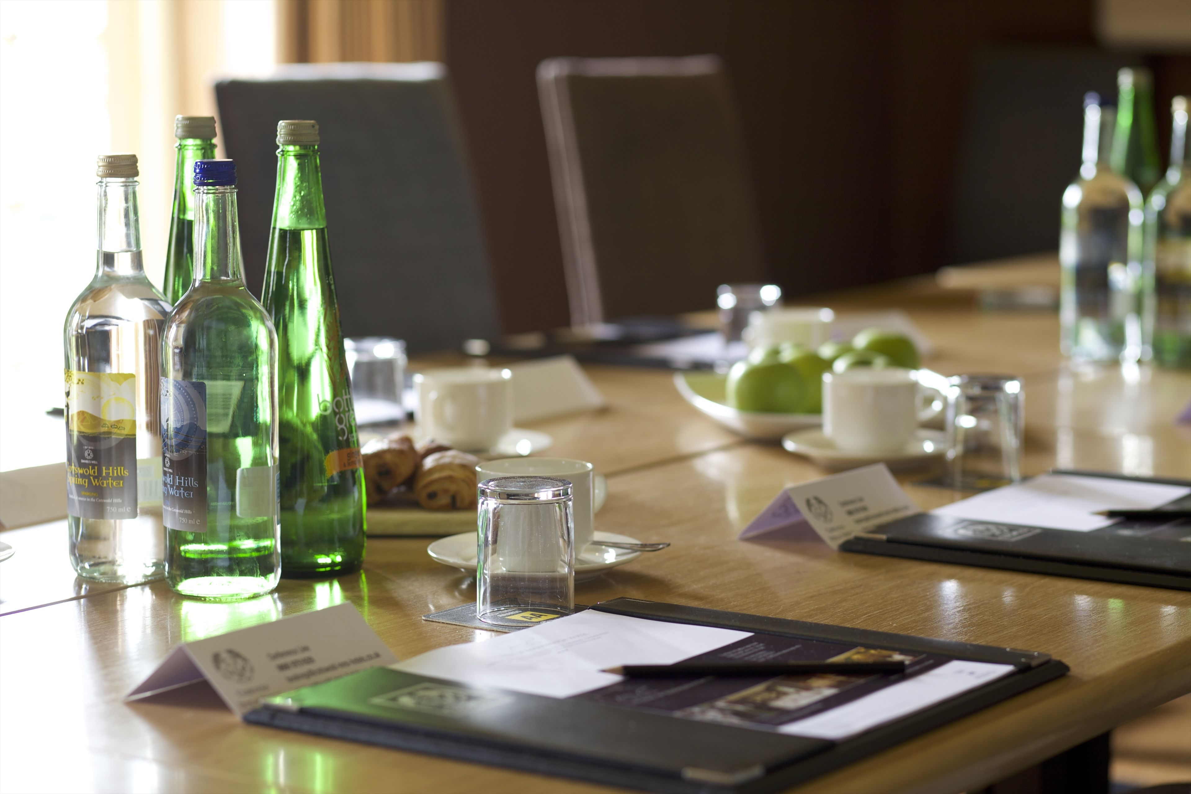A conference table with green glass bottles, a glass of water, a cup, and a plate with croissants.