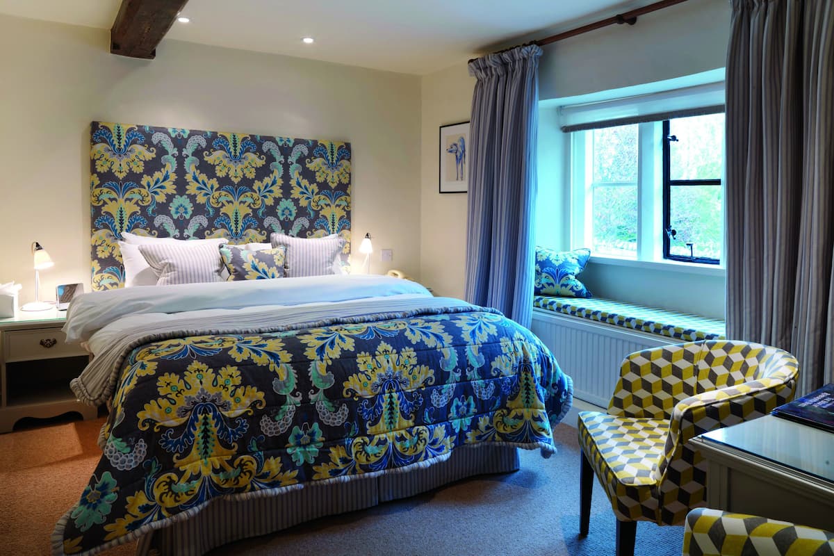 A bedroom with a large bed, a bedside table with a lamp, a window with curtains, and a chair with a patterned cushion.