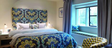A bedroom with a large bed, a bedside table with a lamp, a window with curtains, and a chair with a patterned cushion.