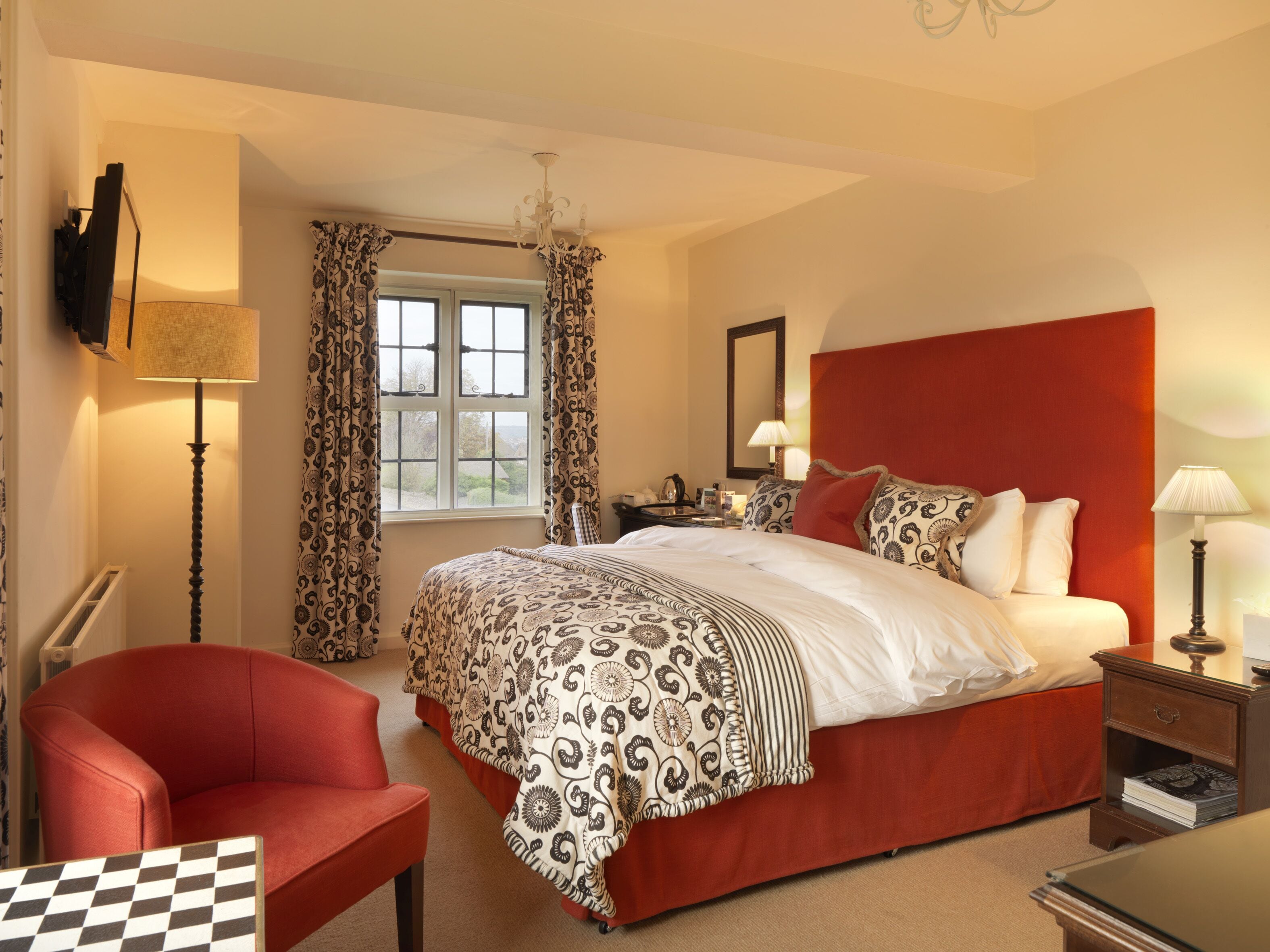 A hotel room with a large bed, a red headboard, a red armchair, a checkered table, and a window with curtains.