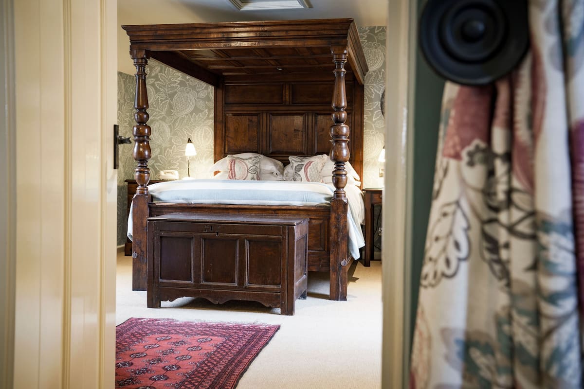 A traditional four-poster bed with a wooden frame, a matching nightstand, and a red patterned rug on the floor.
