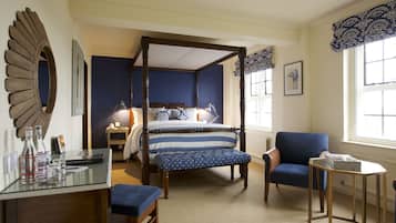 Una cama con dosel con un edredón azul y blanco a rayas, un sillón azul, una mesita redonda con una pila de revistas y un espejo en la pared.
