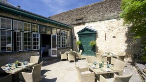 An outdoor patio with wicker furniture and stone flooring, a stone building with a green door, and a clear blue sky. - Bay Tree Hotel (Burford)