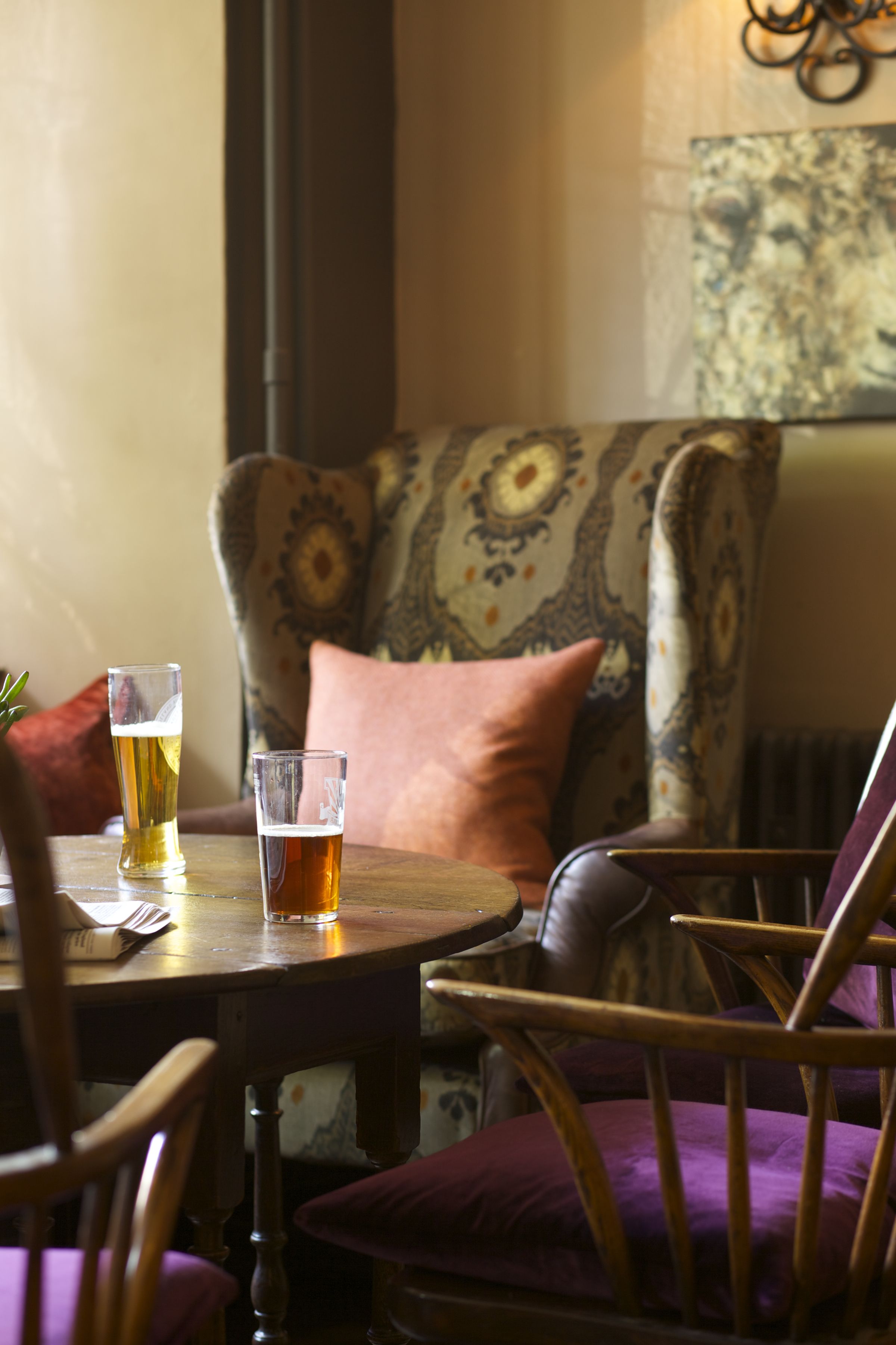 Una mesa redonda con dos vasos de cerveza, una silla con un cojín estampado y una pared decorada con una obra de arte con textura.