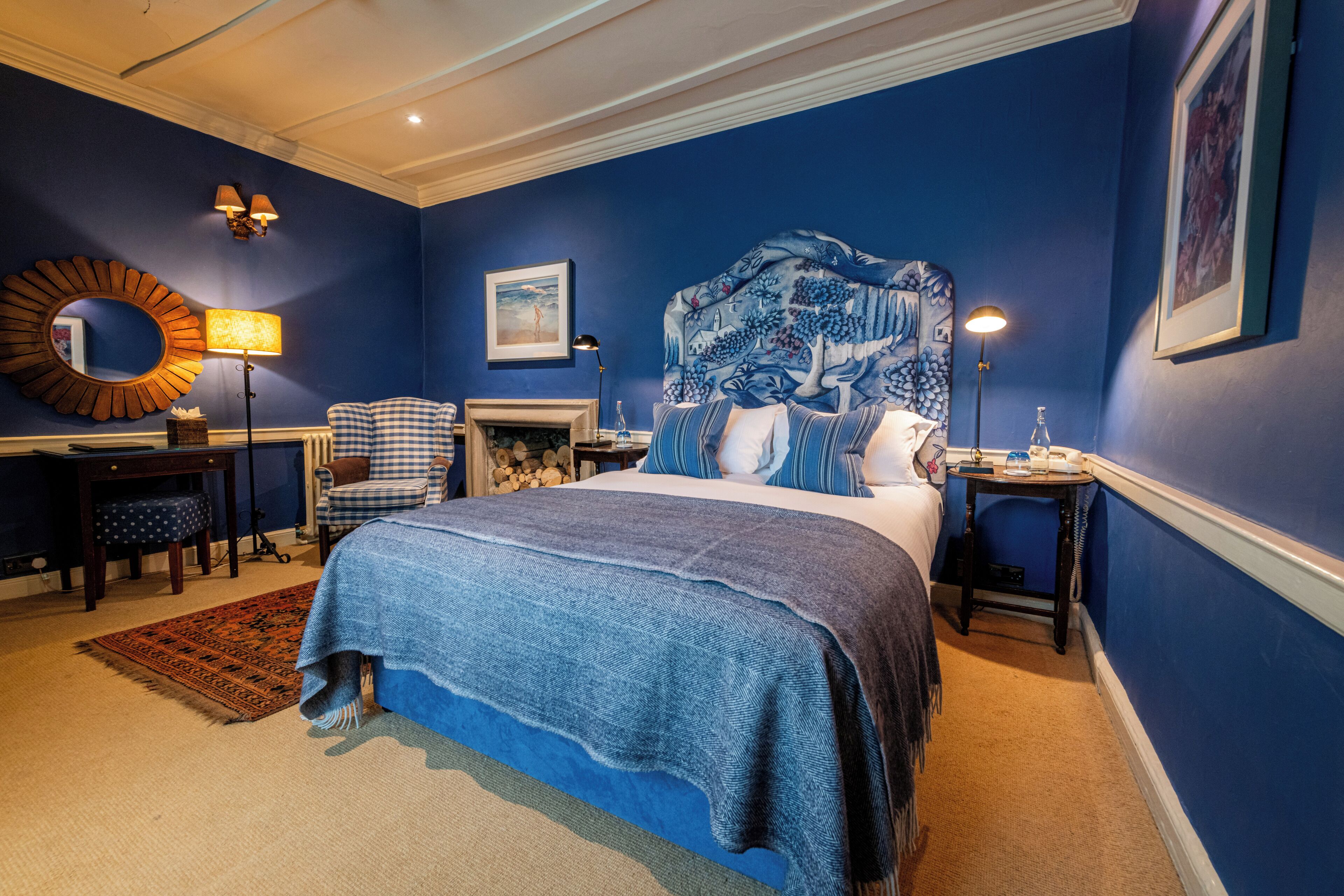 A bedroom with a large bed, a headboard with a floral pattern, a side table with a lamp, a chair, and a fireplace.