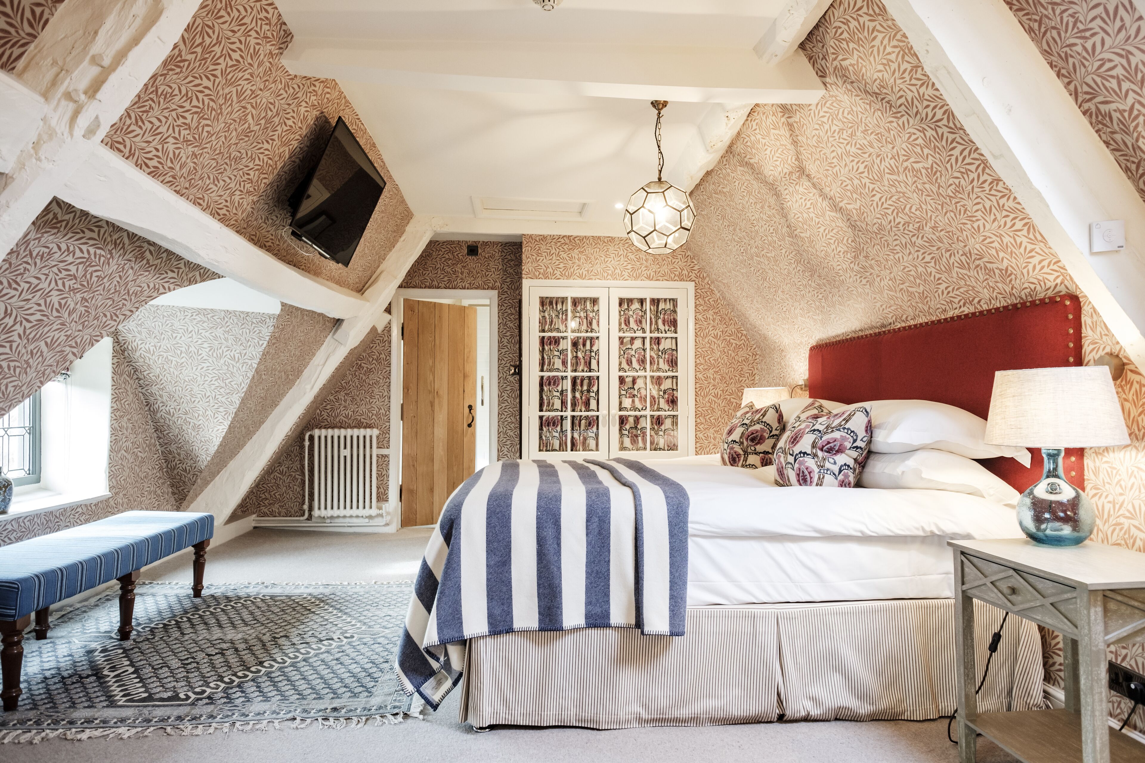 Ein Schlafzimmer mit schrägem Dach, einem Bett mit gestreifter Decke, einem Nachttisch mit Lampe und einem gemusterten Teppich.