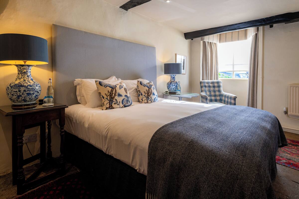 Quarto de hotel com uma cama grande, duas mesas de cabeceira com luminárias, uma cadeira e uma janela com cortinas.
