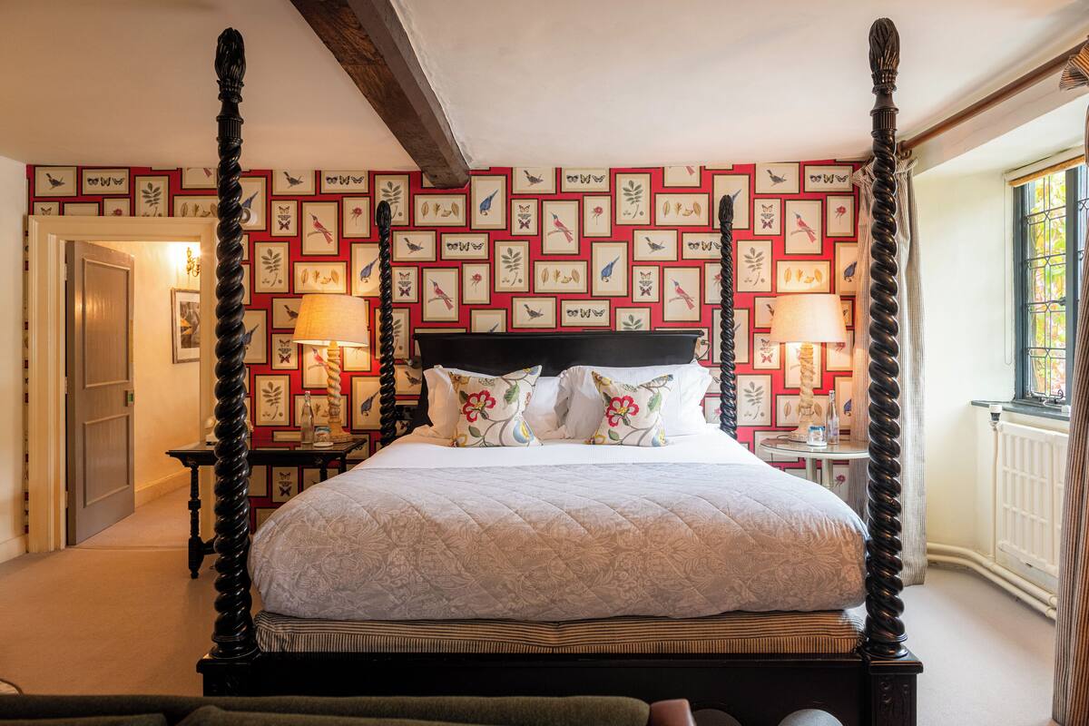 Uma cama com dossel coberta por uma colcha estampada com flores, com duas mesas de cabeceira com abajures de cada lado, em um quarto com parede estampada em vermelho e branco.