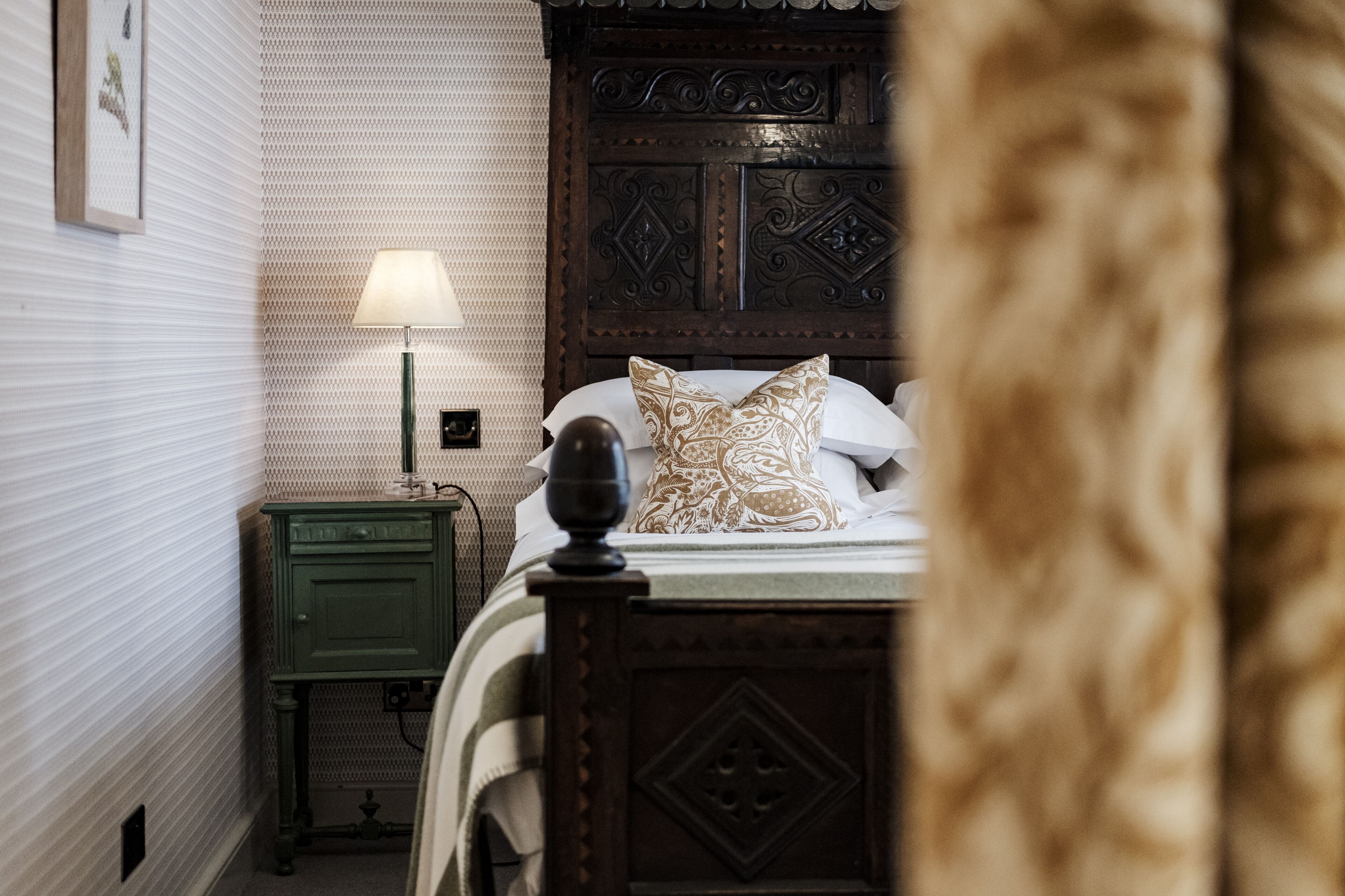 A neatly made bed with a patterned pillow, a bedside lamp, and a green nightstand in a room with patterned wallpaper and a wooden headboard.