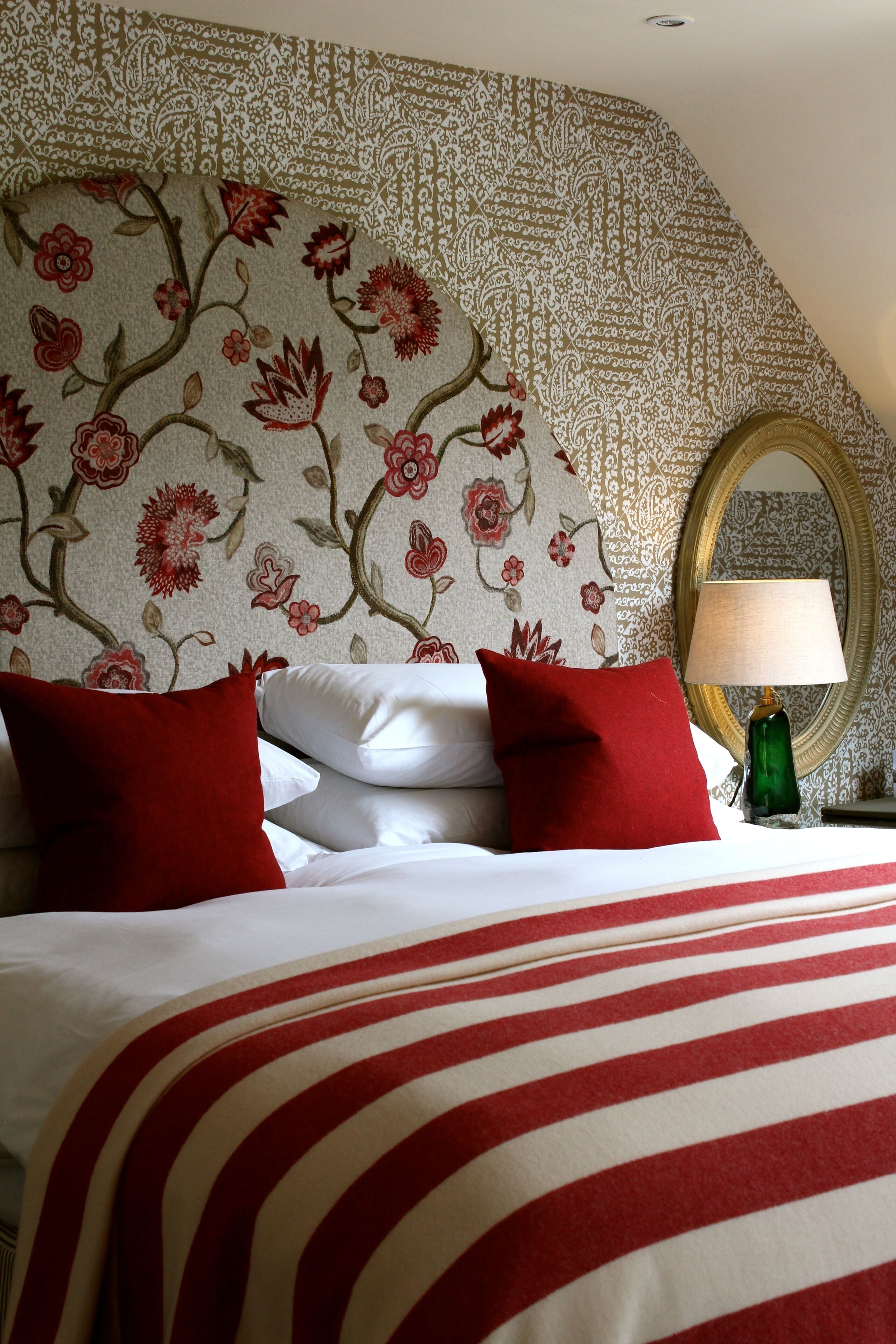 Una habitación de hotel con un cabecero estampado con motivos florales, una cama con ropa de cama roja y blanca, y una lámpara verde sobre una mesita de noche.