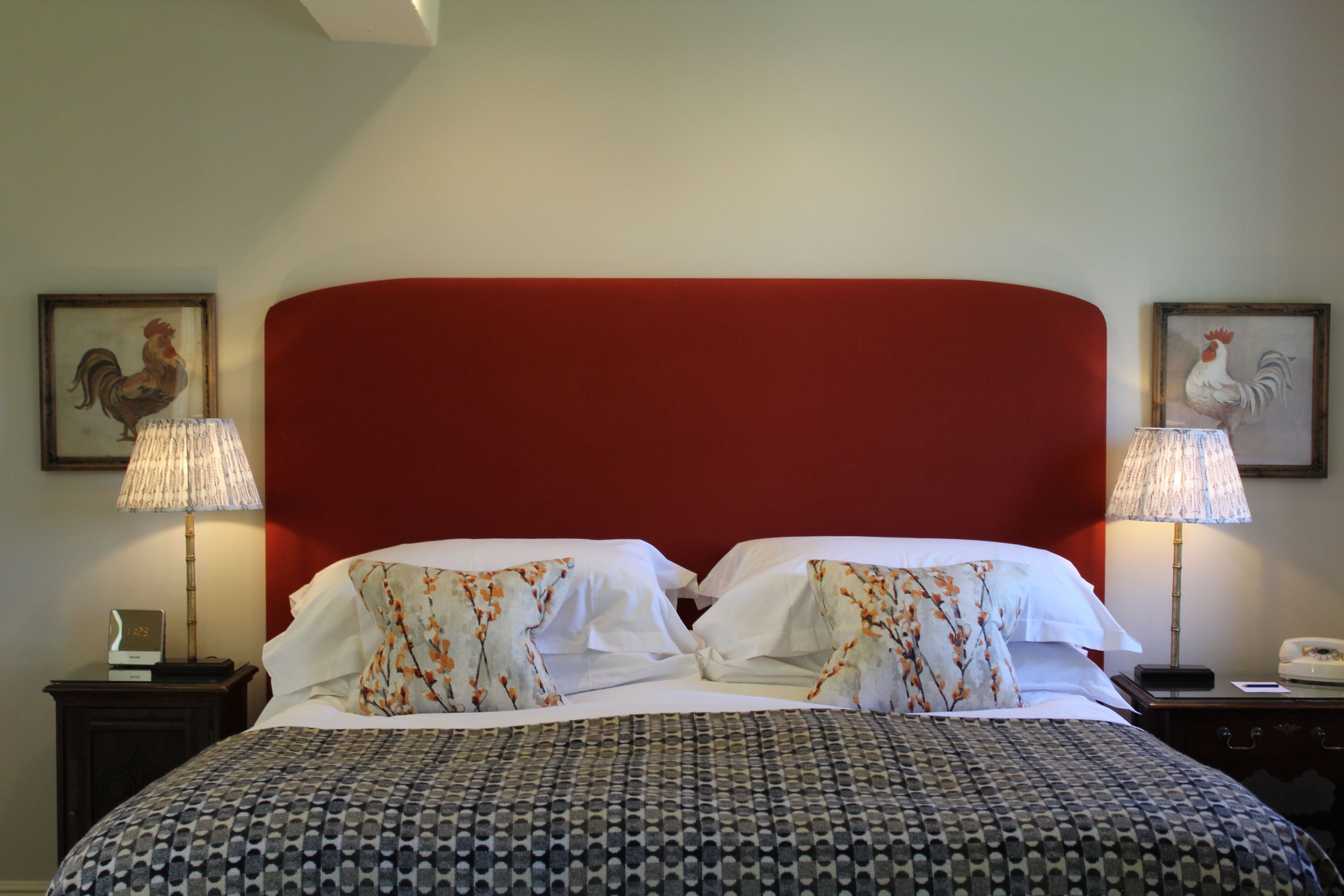 A hotel room with a red headboard, two wall-mounted lamps, a bed with patterned bedding, and two framed rooster pictures on the walls.