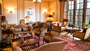 A well-furnished living room with a sofa, armchairs, a coffee table, and a dining table. There are wall sconces, a chandelier, and a large window with curtains. - Cringletie House Hotel (Peebles)