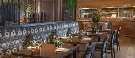 Un restaurante con mesas y bancos de madera, una cocina al fondo y plantas colgantes.