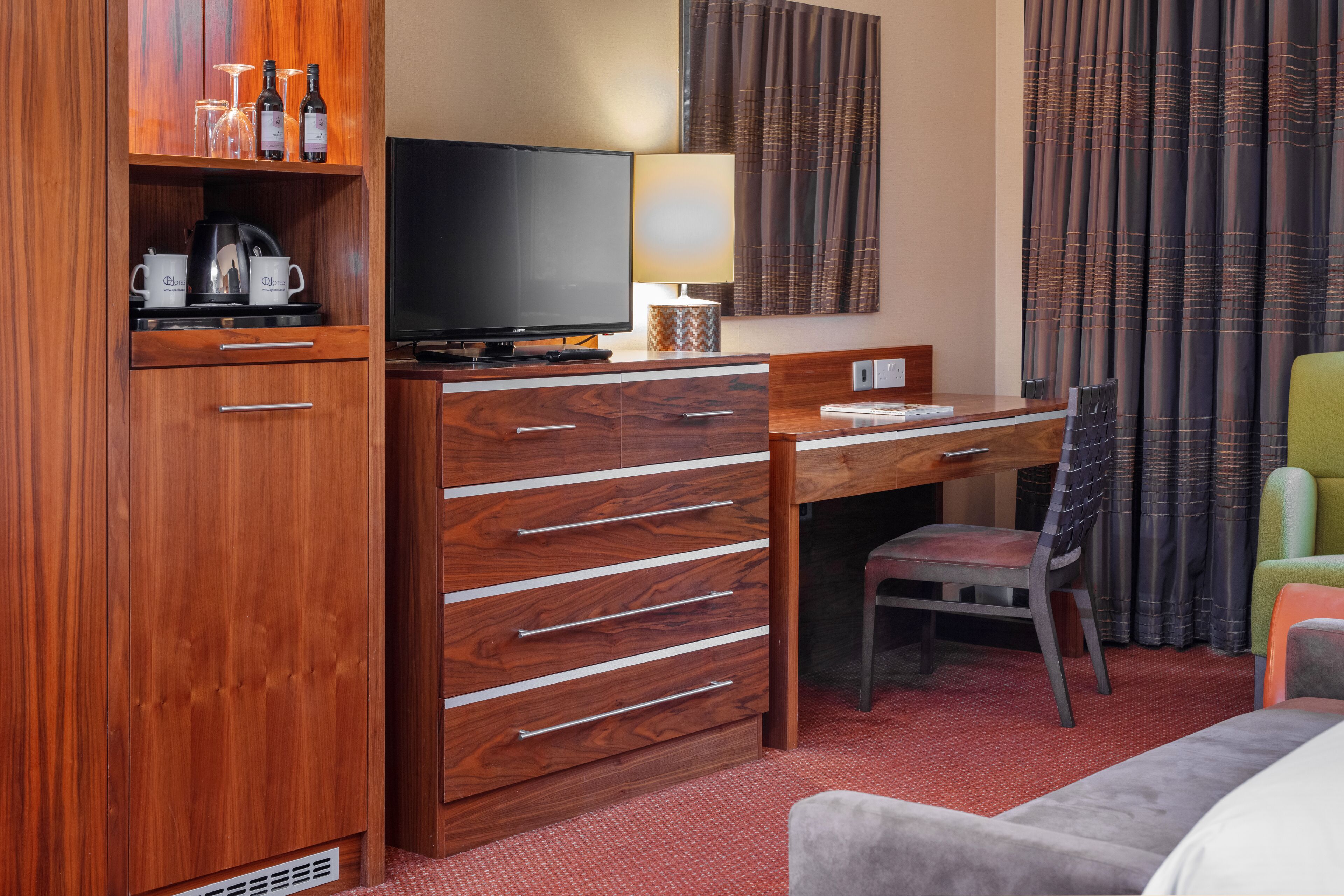 A hotel room with a wooden desk, a television, a lamp, a chair, and a wardrobe.