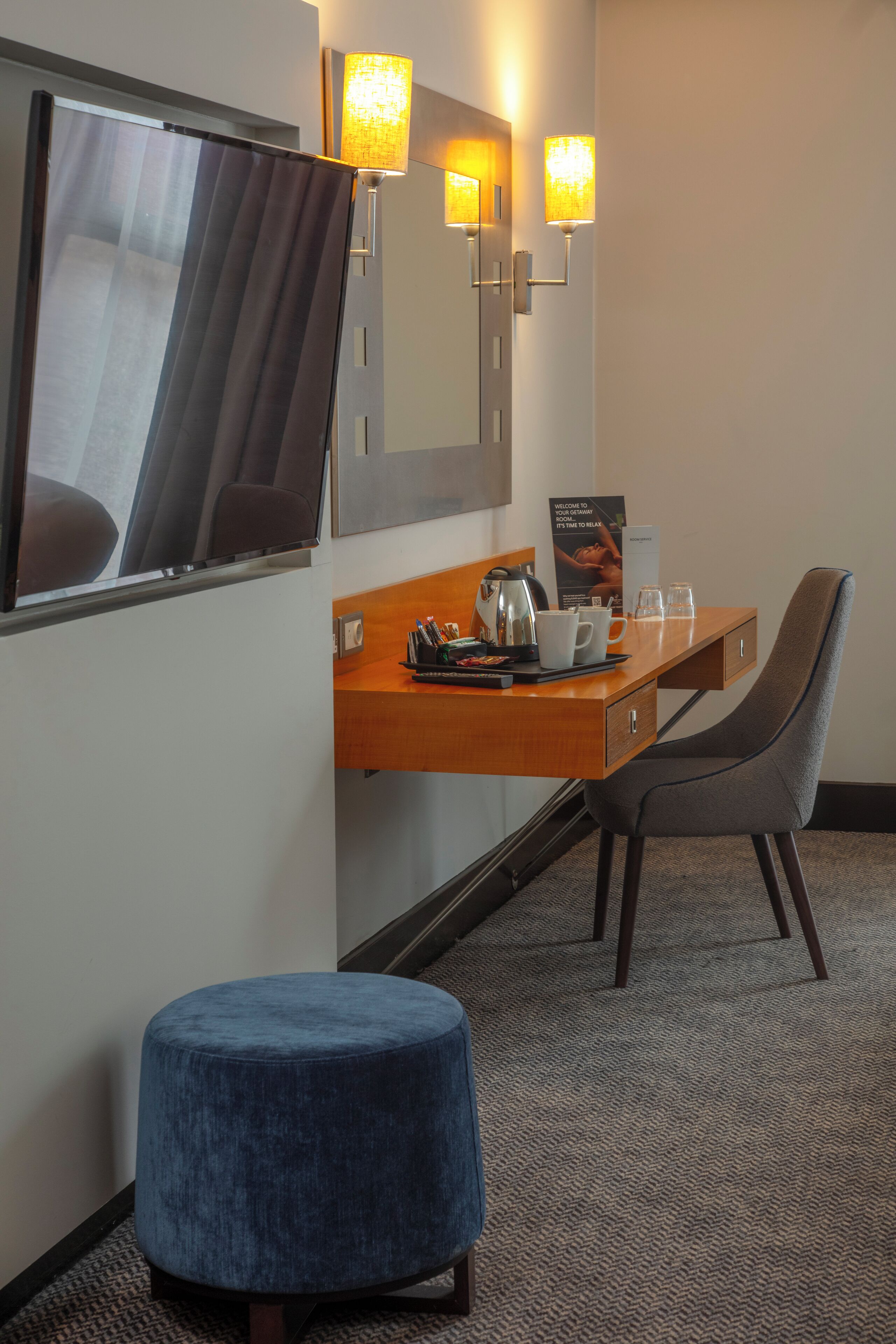 A hotel room with a wooden desk, a chair, a blue ottoman, a mirror, and two wall-mounted lamps.