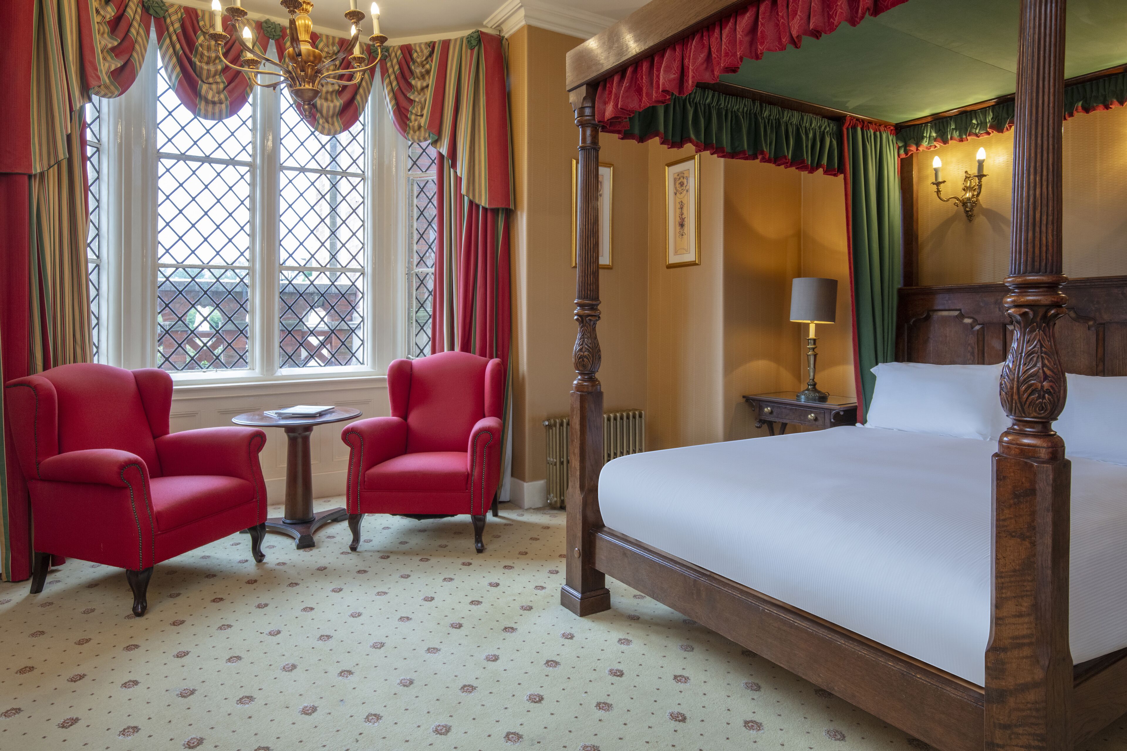 Une chambre à coucher de style traditionnel, avec un lit à baldaquin, deux fauteuils rouges, une table ronde et une fenêtre avec des rideaux.