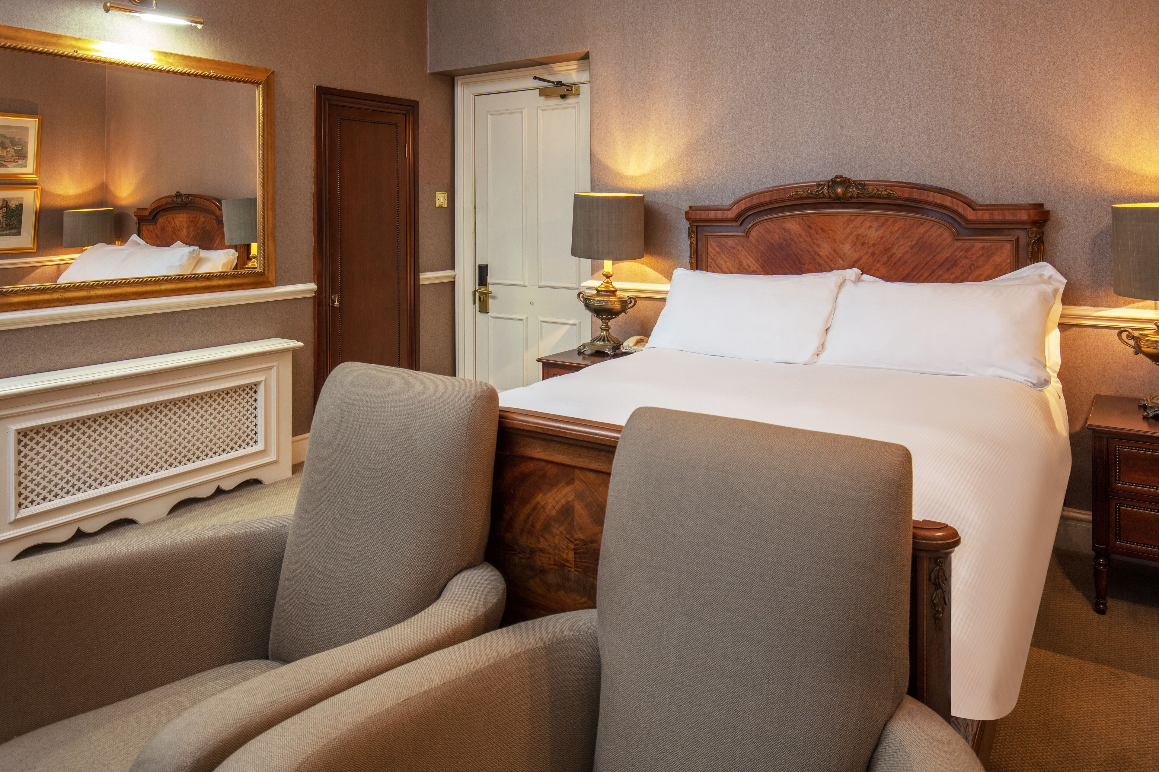 A hotel room with a large bed, two armchairs, a wooden headboard, a nightstand with a lamp, and a mirror above the bed.