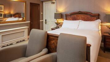 A hotel room with a large bed, two armchairs, a wooden headboard, a nightstand with a lamp, and a mirror above the bed.