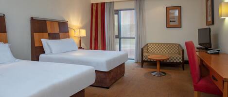 A hotel room with two beds, a desk, a chair, a small table, and a television.
