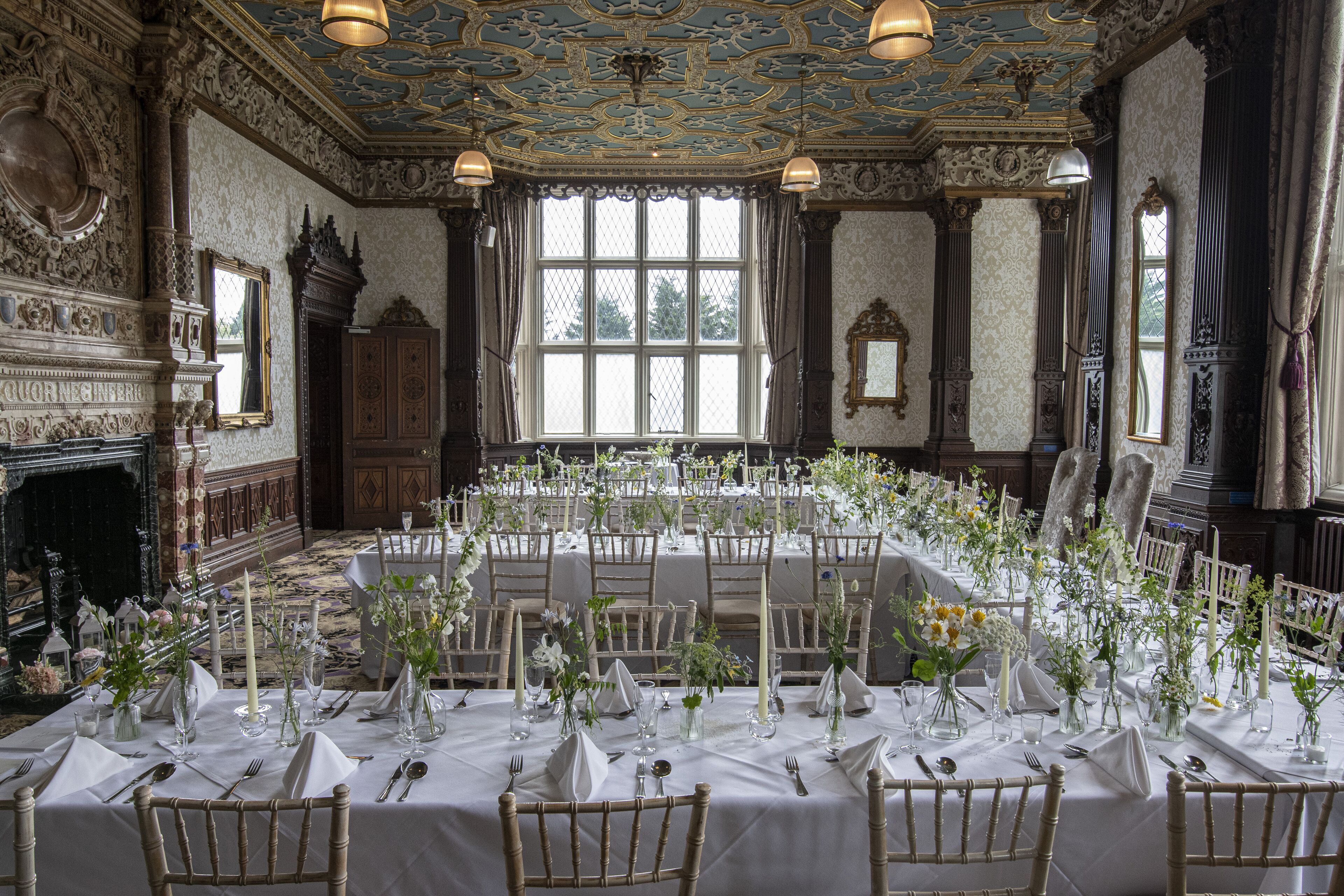 Un salón de banquetes con techo ornamentado, amplias ventanas y chimenea. Las mesas están dispuestas con manteles blancos y centros de mesa florales.