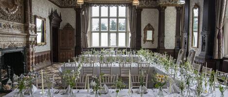 Un salón de banquetes con techo ornamentado, amplias ventanas y chimenea. Las mesas están dispuestas con manteles blancos y centros de mesa florales.