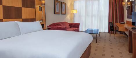 A hotel room with a large bed, a desk, a chair, a red sofa, and a TV.
