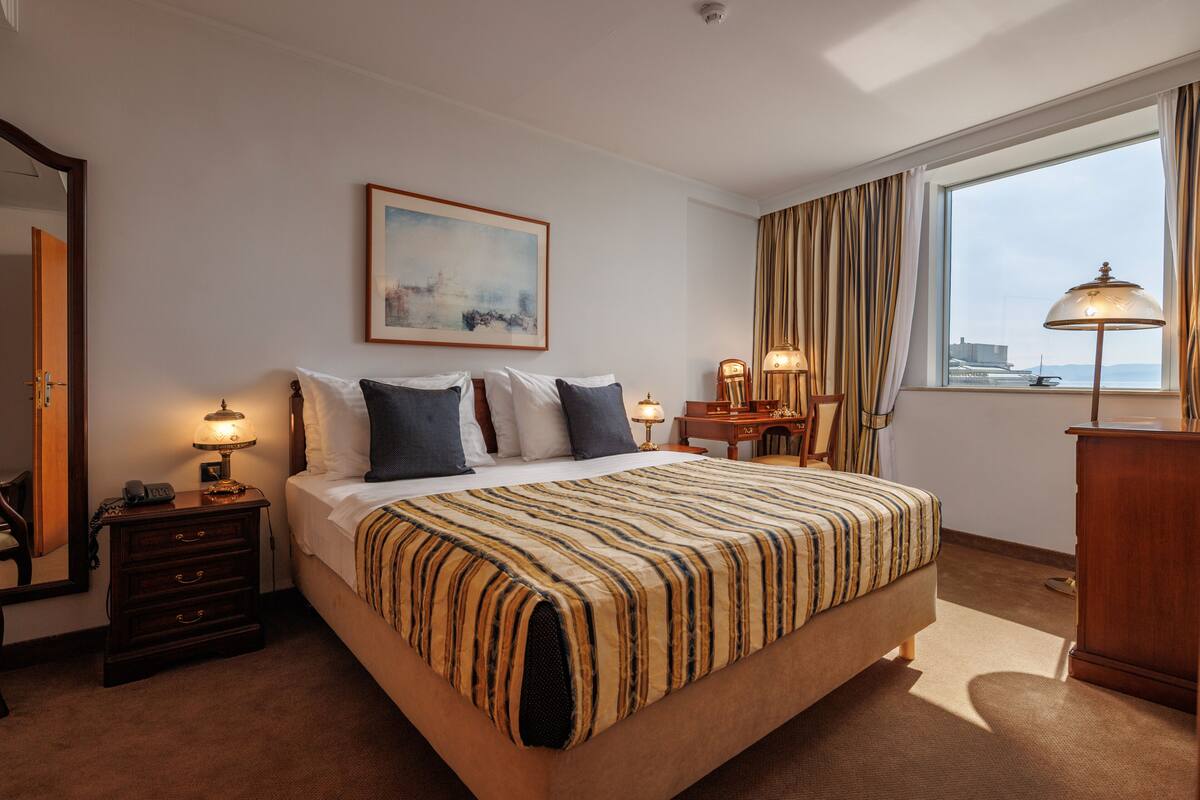Habitación de hotel con una cama grande, una mesita de noche con lámpara, un escritorio, una silla, un armario y un ventanal con cortinas.