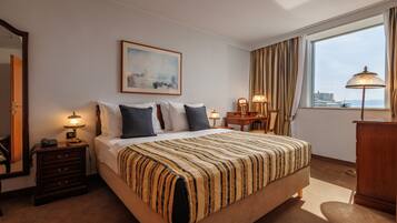 A hotel room with a large bed, a bedside table with a lamp, a desk, a chair, a wardrobe, and a large window with curtains.