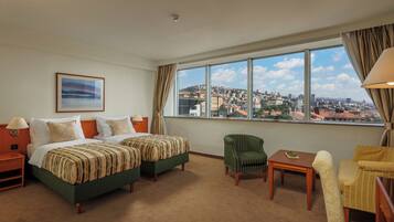 Habitación de hotel con dos camas, vistas a la ciudad, un sillón verde, un sillón amarillo y una mesita.