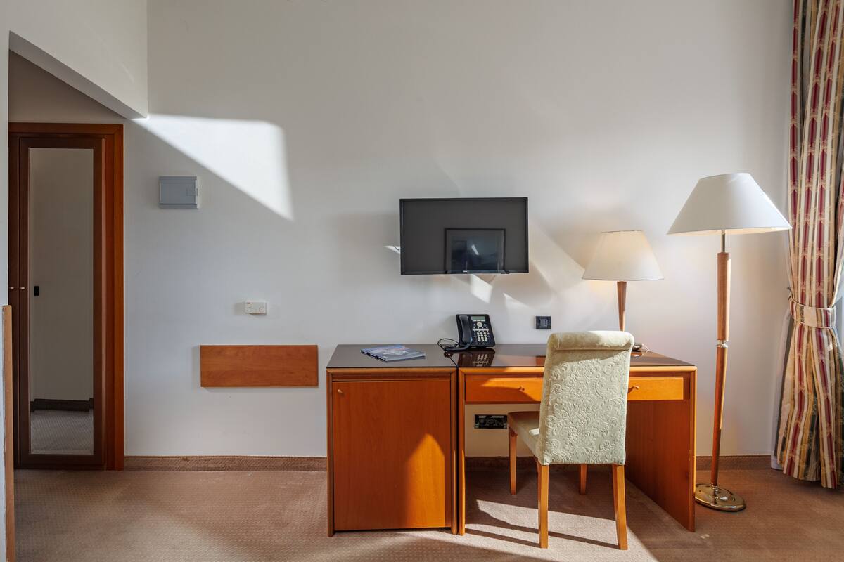 Una habitación de hotel con un escritorio de madera, una silla, un televisor de pantalla plana montado en la pared, una lámpara de escritorio y una ventana con cortinas.