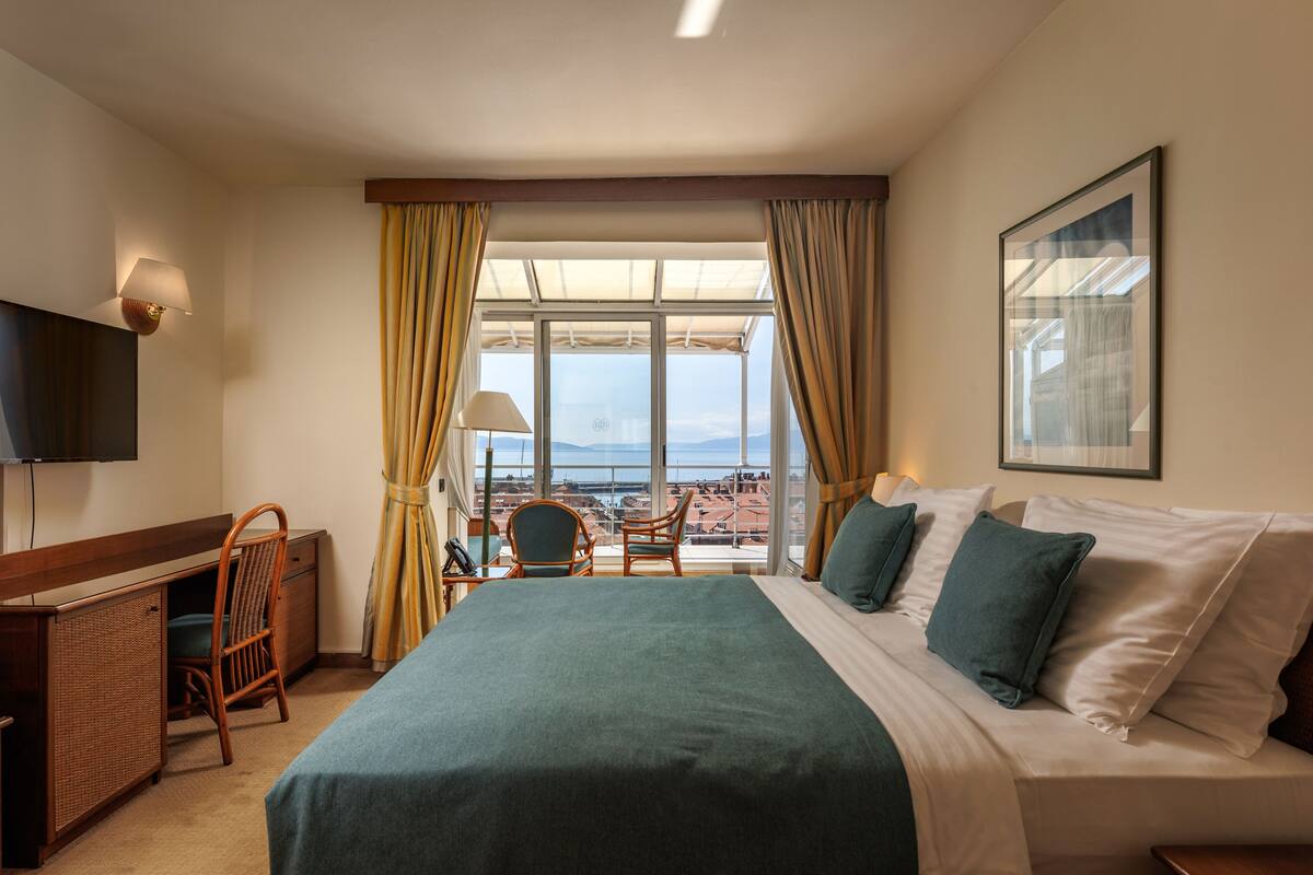 Una habitación de hotel con cama, escritorio, silla, televisión y un ventanal con cortinas.