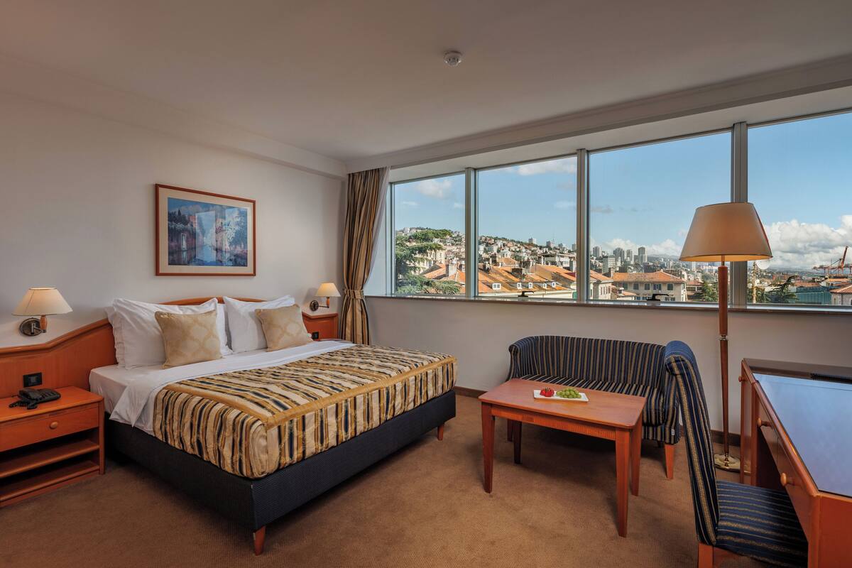 Habitación de hotel con una cama grande, un escritorio, una silla y vistas a un paisaje urbano a través de amplias ventanas.
