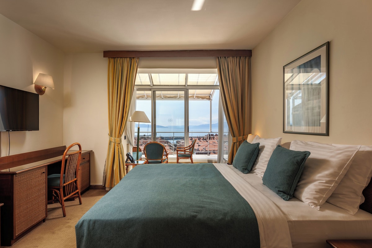 Una habitación de hotel con cama, escritorio, silla, televisión y un ventanal con cortinas.