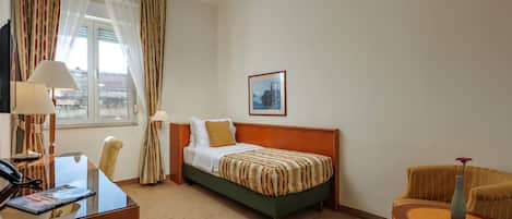 Una habitación de hotel con una cama, un escritorio con teléfono, una silla, una mesita con un florero y una ventana con cortinas.