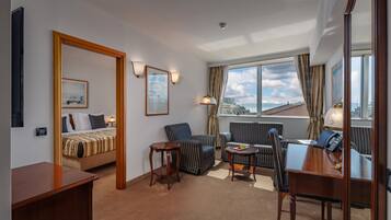 A hotel room with a bed, two armchairs, a small table, a desk, and a large window with a view of the city.