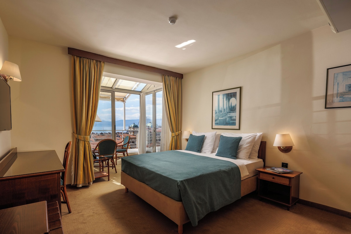 A hotel room with a bed, desk, chair, and a view of the city through large windows.