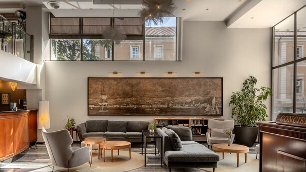A modern hotel lobby with a reception desk, seating area, large artwork, and a view of the city through large windows.