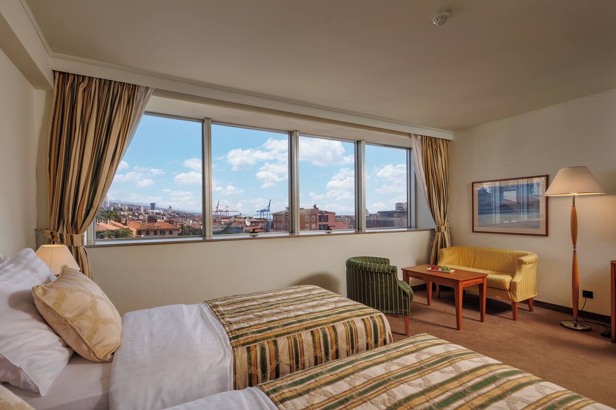 Una habitación de hotel con un ventanal grande, una cama, una silla, una mesa y una lámpara.