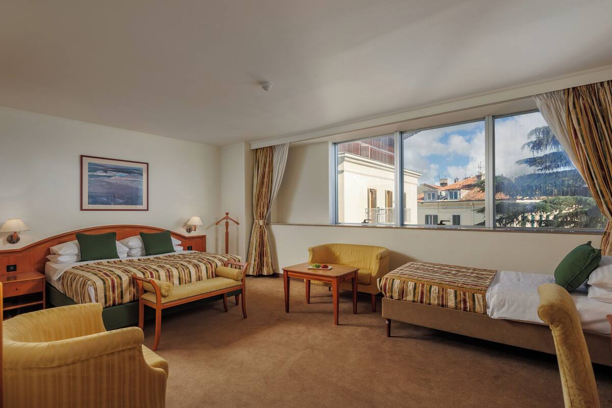 Habitación de hotel con cama, dos sillas, una mesita y un ventanal grande que ofrece vistas a edificios y árboles.