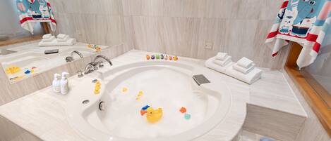 A bathroom with a large bathtub, a mirror, and a sink. There are towels and slippers on the floor.