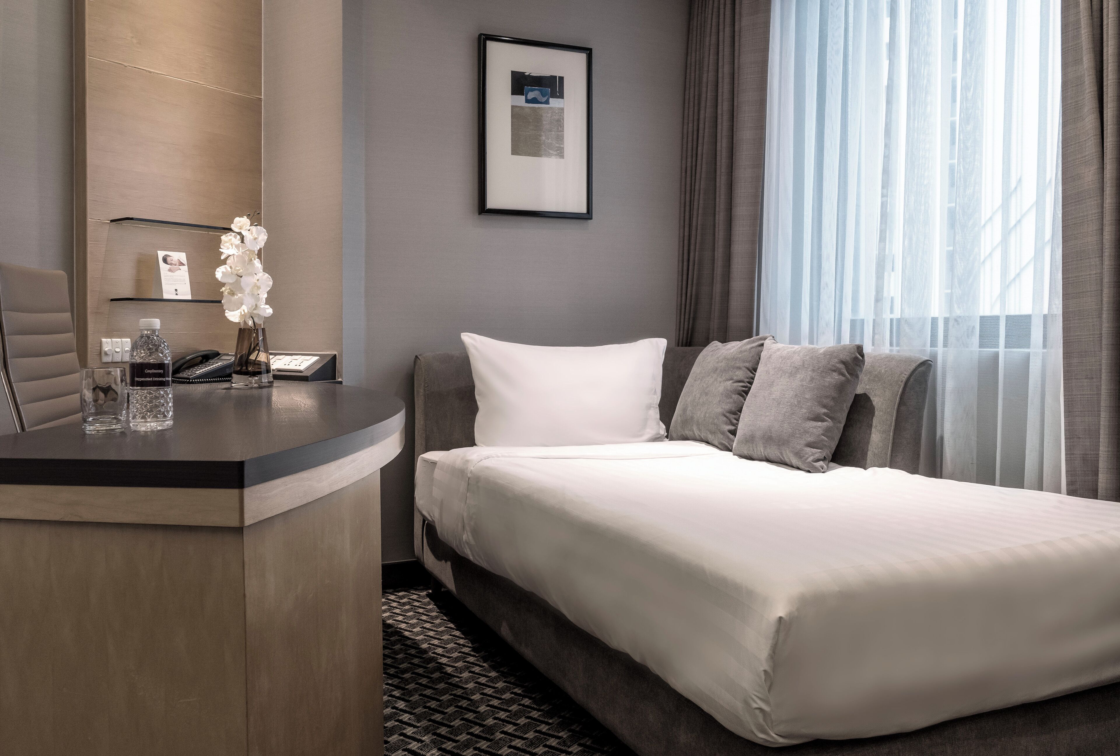 A hotel room with a bed, a desk with a phone, a vase with flowers, and a framed picture on the wall.