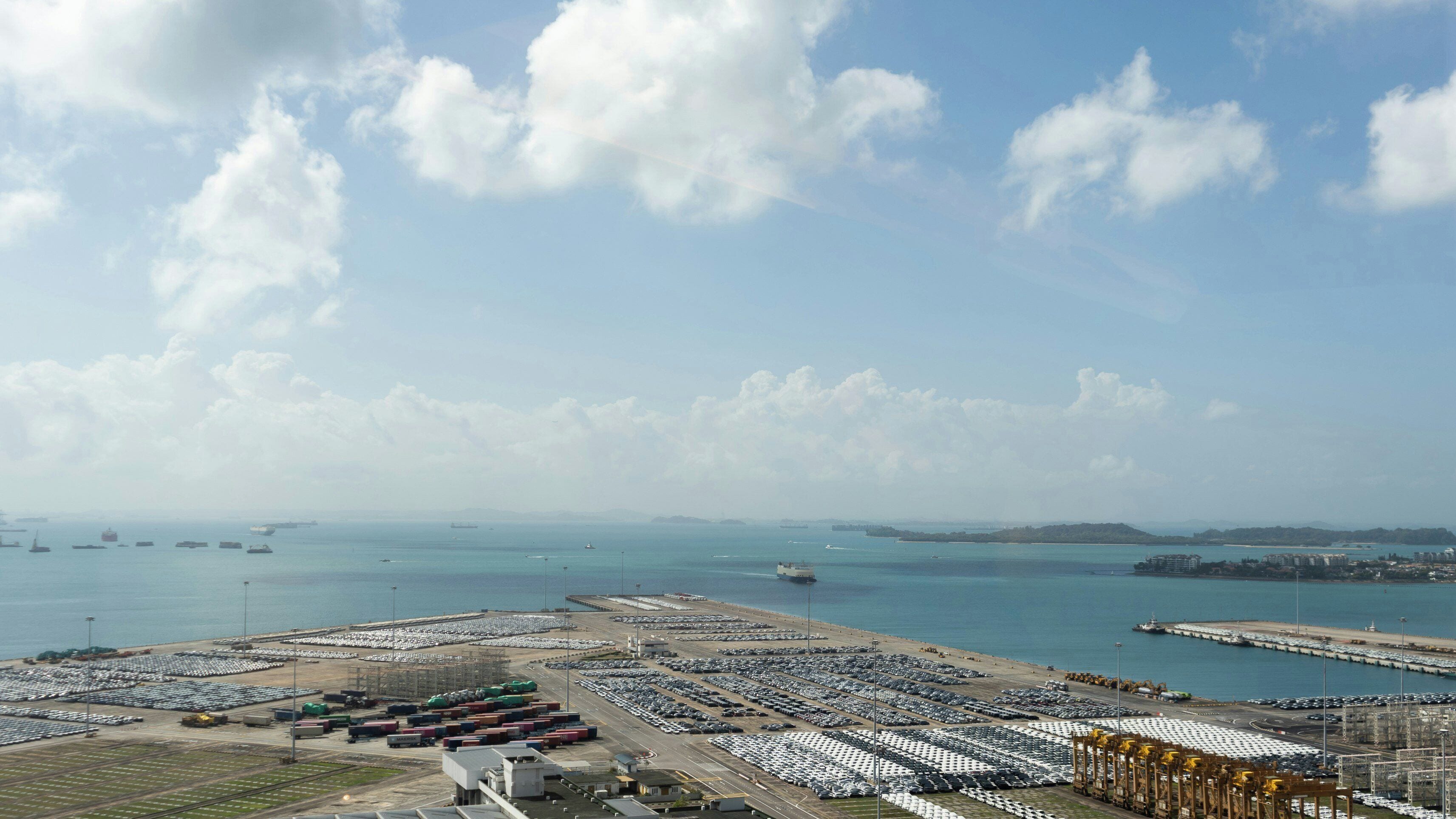 A large parking area with rows of parked cars and a port in the background.