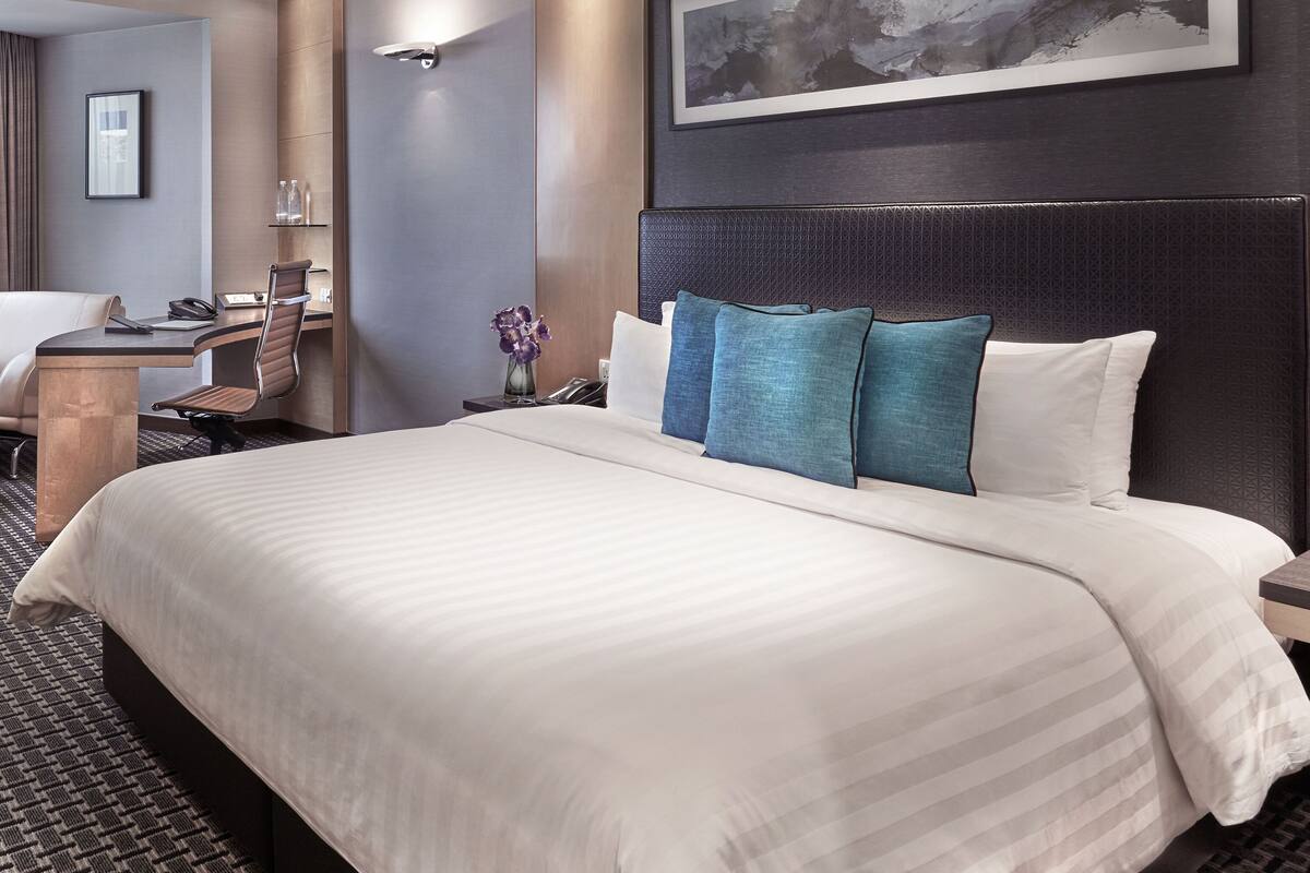 Une chambre d’hôtel avec un grand lit, un bureau avec une chaise, une petite table avec un vase de fleurs et une horloge fixée au mur.