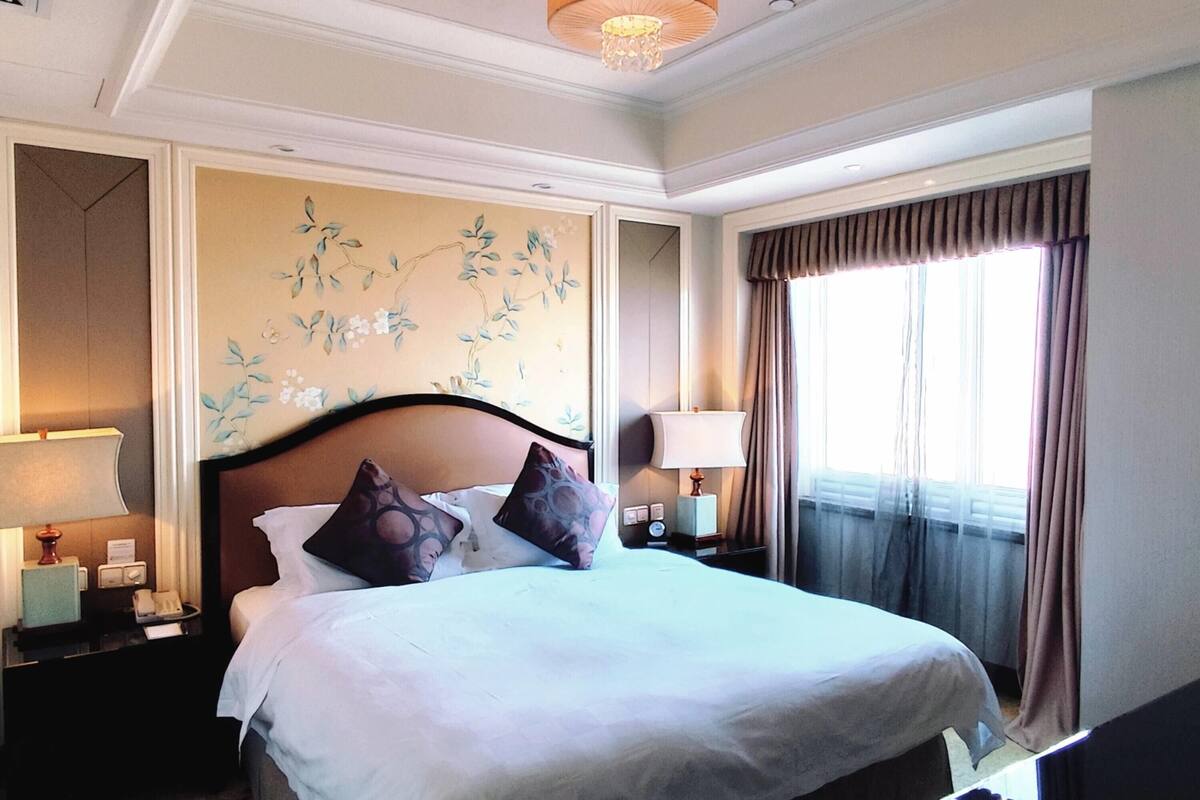Habitación de hotel con una cama grande, dos mesitas de noche, una lámpara de pared y un ventanal con cortinas.
