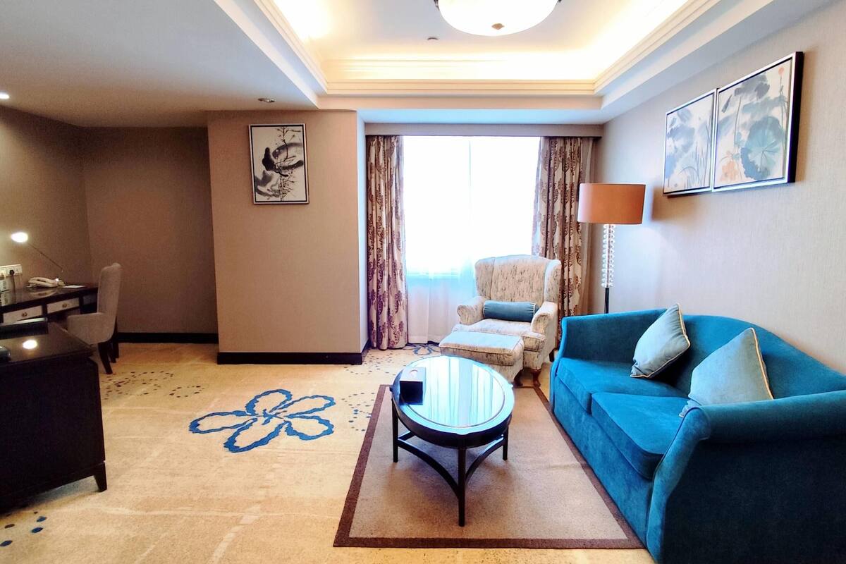Habitación de hotel con sofá, sillón, mesita y un amplio ventanal con cortinas.
