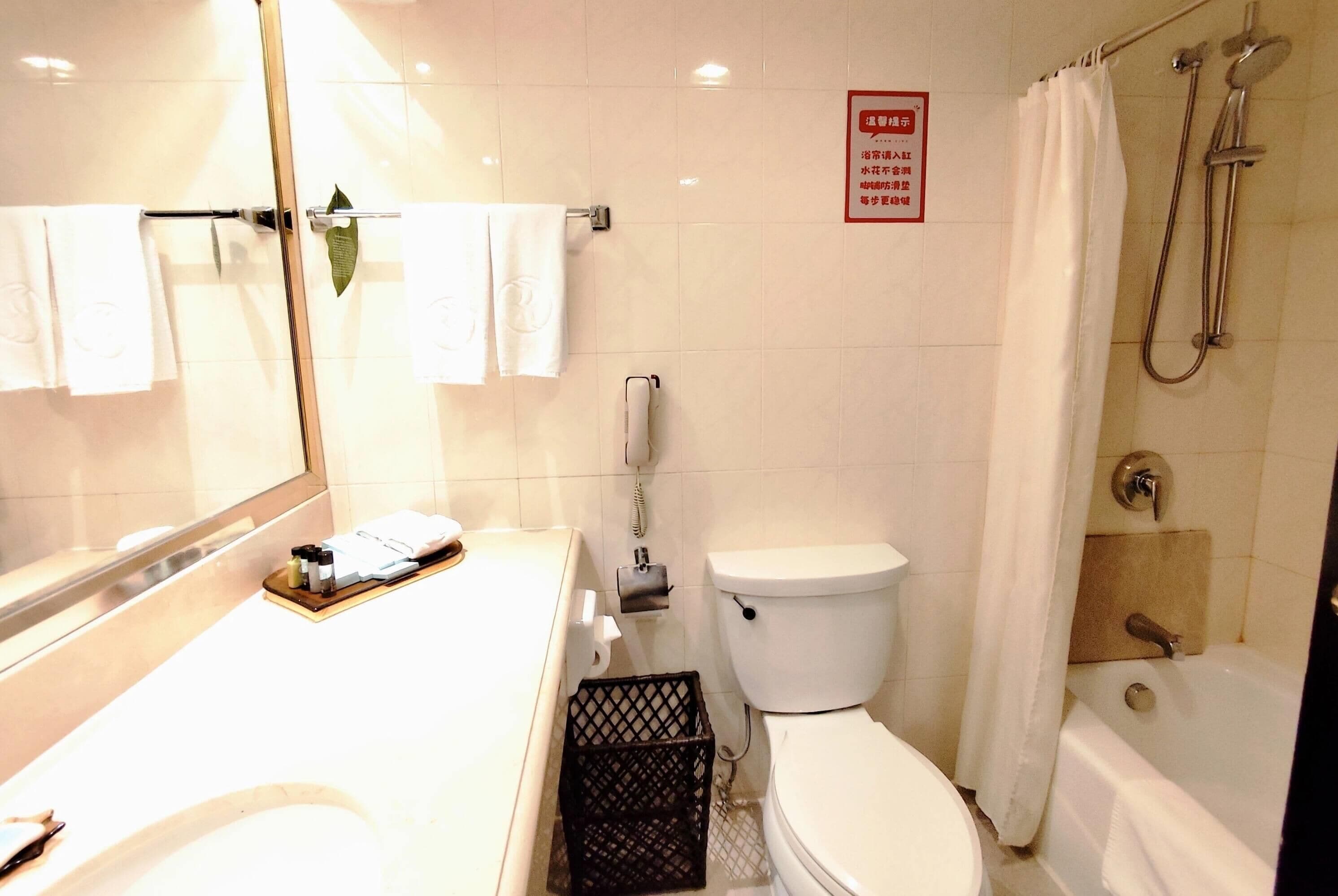 Een badkamer met toilet, wastafel en douche. Er hangt een bord aan de muur en er staat een mandje bij de wastafel.