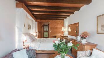 A cozy bedroom with a wooden ceiling, a bed with white bedding, a bedside table with a lamp, a dresser, and a floral arrangement on the dresser.