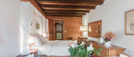 Un dormitorio acogedor con techo de madera, una cama con ropa de cama blanca, una mesita de noche con lámpara, un aparador y un arreglo floral sobre el aparador.